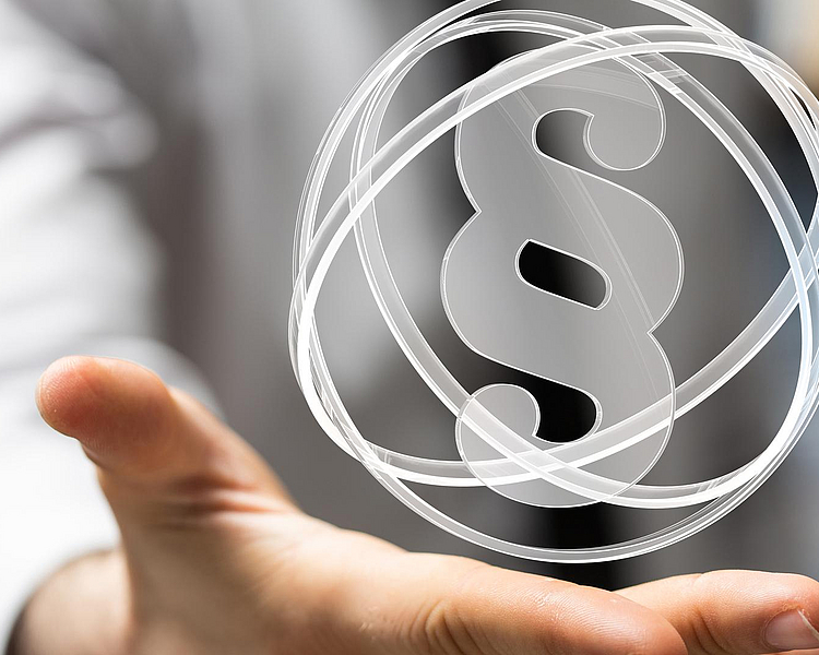 Eine Hand hält ein schwebendes Paragraphen-Symbol, das in einem transparenten Kreis dargestellt ist. Das Bild vermittelt ein Gefühl von rechtlichen Themen oder juristischer Beratung.