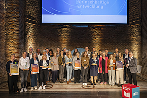 Eine Gruppe von Preisträgern steht auf einer Bühne in einem historischen Raum. Sie halten Auszeichnungen für nachhaltige Entwicklung in den Händen. Im Hintergrund ist ein Bildschirm mit dem Schriftzug "für nachhaltige Entwicklung" zu sehen.