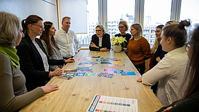Eine Gruppe von zehn Personen sitzt oder steht um einen Tisch. Auf dem Tisch liegen bunte Materialien und Karten. Einige Personen diskutieren aktiv, während andere aufmerksam zuhören. Im Hintergrund sind große Fenster mit Blick auf die städtische Umgebung sichtbar.