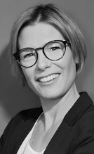 Porträt einer lächelnden Frau mit Kurzhaarschnitt, die eine schwarze Blazerjacke und eine weiße Bluse trägt. Sie steht mit verschränkten Armen vor einem neutralen Hintergrund und vermittelt Selbstbewusstsein und Professionalität.