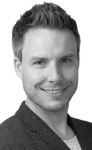 Ein lächelnder Mann mit kurzen, hellen Haaren und einem gepflegten Bart trägt ein dunkles Blazerjackett und ein helles T-Shirt. Der Hintergrund ist neutral und hell, was den Fokus auf sein freundliches Gesicht lenkt.