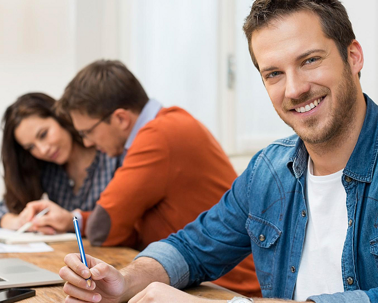 Ein lächelnder Mann in einem blauen Hemd sitzt an einem Tisch und hält einen Stift in der Hand. Im Hintergrund sind zwei Personen, die zusammenarbeiten. Auf dem Tisch liegen Papierblätter und ein Laptop ist sichtbar.