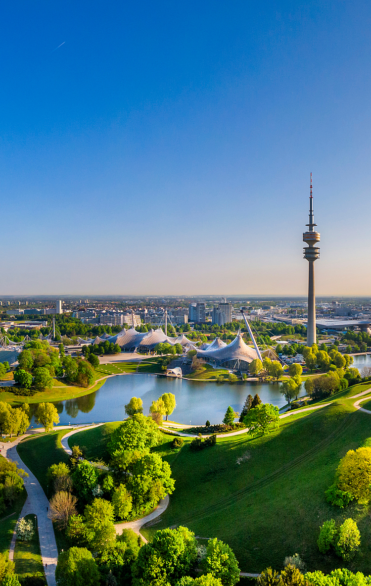 Ein Panoramablick auf eine grüne Landschaft mit einem Teich, umgeben von Bäumen und gepflegten Parkanlagen. Im Hintergrund ist der Fernsehturm zu sehen, während die Sonne am Horizont aufgeht und den Himmel erhellt.
