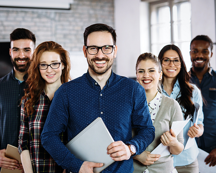 Eine Gruppe von sechs lächelnden Personen steht in einem modernen Büro. Sie tragen unterschiedliche Outfits, halten Dokumente oder Laptops in den Händen und wirken freundlich und motiviert. Die Szene vermittelt ein Gefühl von Teamarbeit und Zusammenarbeit.