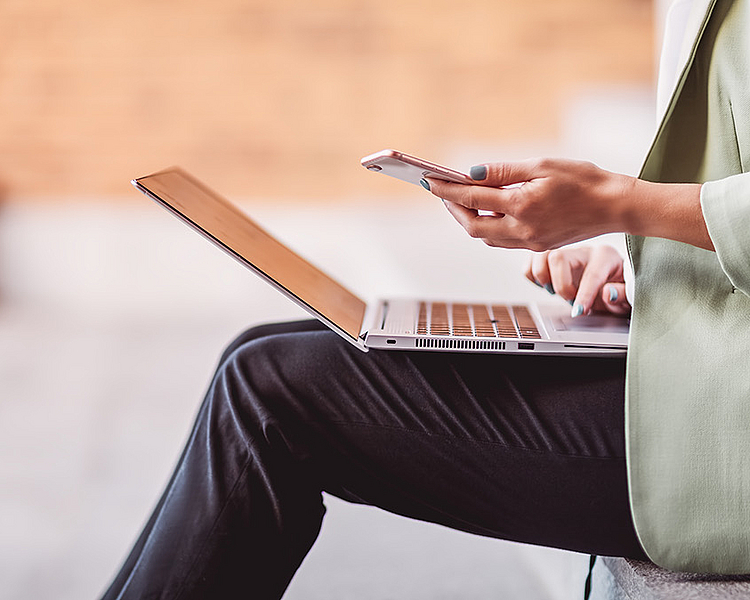Eine Person sitzt auf einer Treppe und hält ein Smartphone in der Hand, während sie auf einem Laptop vor sich tippt. Die Person trägt einen Blazer und wirkt in einer entspannten Arbeitsumgebung.