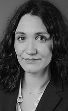 Porträt einer Frau mit langen, lockigen Haaren, die in einem dunklen Blazer gekleidet ist. Sie schaut direkt in die Kamera und hat einen neutralen Gesichtsausdruck. Der Hintergrund ist einfarbig und unscharf.