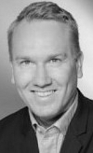 Porträt einer Person mit kurzen, hellen Haaren, die in einem schwarzen Anzug und einem hellen Hemd gekleidet ist. Die Person lächelt freundlich und ist vor einem neutralen Hintergrund abgebildet.