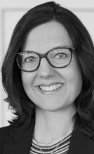 Porträt einer Frau mit langen, dunklen Haaren und einer Brille. Sie trägt einen Blazer und hat ein freundliches Lächeln. Der Hintergrund ist unscharf und neutral gestaltet.