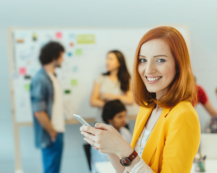 Eine lächelnde Frau mit rotem Haar, die ein Handy in der Hand hält, steht im Vordergrund. Im Hintergrund sind mehrere Personen in einem kreativen Arbeitsumfeld zu sehen, die an einem Whiteboard mit Notizen arbeiten.