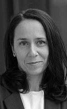 Eine Frau mit langen, dunklen Haaren lächelt leicht in die Kamera. Sie trägt ein schwarzes Blazerjackett und hat eine neutrale Umgebung im Hintergrund. Die Aufnahme ist in Schwarz-Weiß gehalten und vermittelt einen professionellen Eindruck.