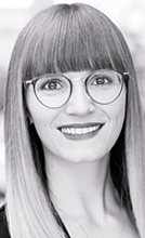 Ein Porträt einer Person mit langen, blonden Haaren und Brille. Sie lächelt freundlich und schaut direkt in die Kamera. Der Hintergrund ist unscharf, was den Fokus auf das Gesicht der Person legt.