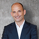 Ein lächelnder Mann mit Glatze trägt einen dunklen Anzug und ein weißes Hemd. Er steht vor einem neutralen, grauen Hintergrund und hat eine freundliche Ausstrahlung.