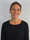 Porträt einer Frau mit langen, braunen Haaren, die lächelt und in einem schwarzen Oberteil gekleidet ist. Sie steht vor einem hellen Hintergrund und wirkt freundlich und zugänglich.