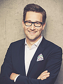 Ein lächelnder Mann in einem schwarzen Anzug mit einem weißen Hemd und einer farbigen Einstecktücher steht mit gekreuzten Armen vor einem neutralen, grauen Hintergrund. Er trägt eine Brille und hat kurzes, gestyltes Haar.