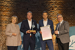 Eine Gruppe von vier Personen steht auf einer Bühne. Zwei Männer halten eine Auszeichnung und einen Preis in den Händen. Die Frau links zeigt lächelnd in die Kamera. Der Hintergrund besteht aus einer Ziegelwand. Die Atmosphäre ist feierlich.