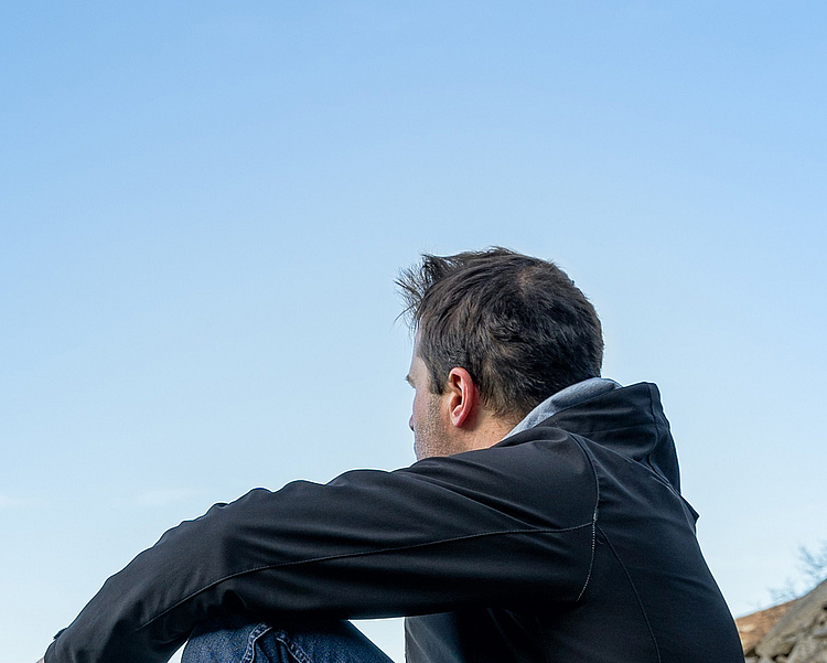 Ein Mann sitzt auf einem großen Stein in den Bergen, umgeben von einer natürlichen Landschaft. Er betrachtet den Horizont und hält eine Tasse in der Hand, während er entspannt die Aussicht genießt. Der Himmel ist klar und blau.