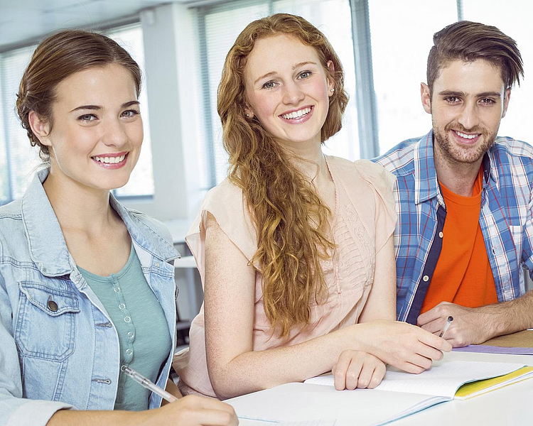 BWV Bildungsverband Duales Studium Vier junge Erwachsene sitzen an einem Tisch in einem hellen Raum. Sie lächeln freundlich in die Kamera. Auf dem Tisch liegen Notizblätter und ein Laptop. Die Atmosphäre wirkt entspannt und gemeinschaftlich, als ob sie zusammenarbeiten oder lernen.