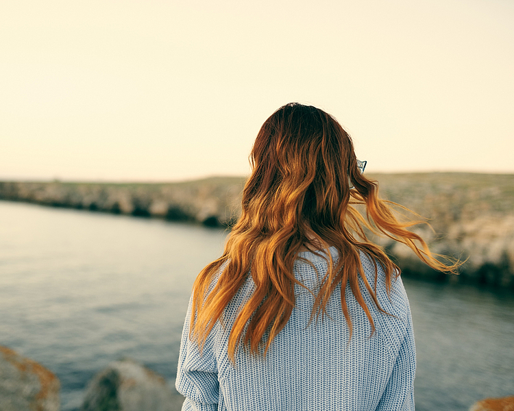 Eine Person mit langen, welligen Haaren steht am Rand eines Gewässers und blickt nach außen. Der Horizont ist sanft im Hintergrund sichtbar, und die Atmosphäre wirkt ruhig und friedlich.