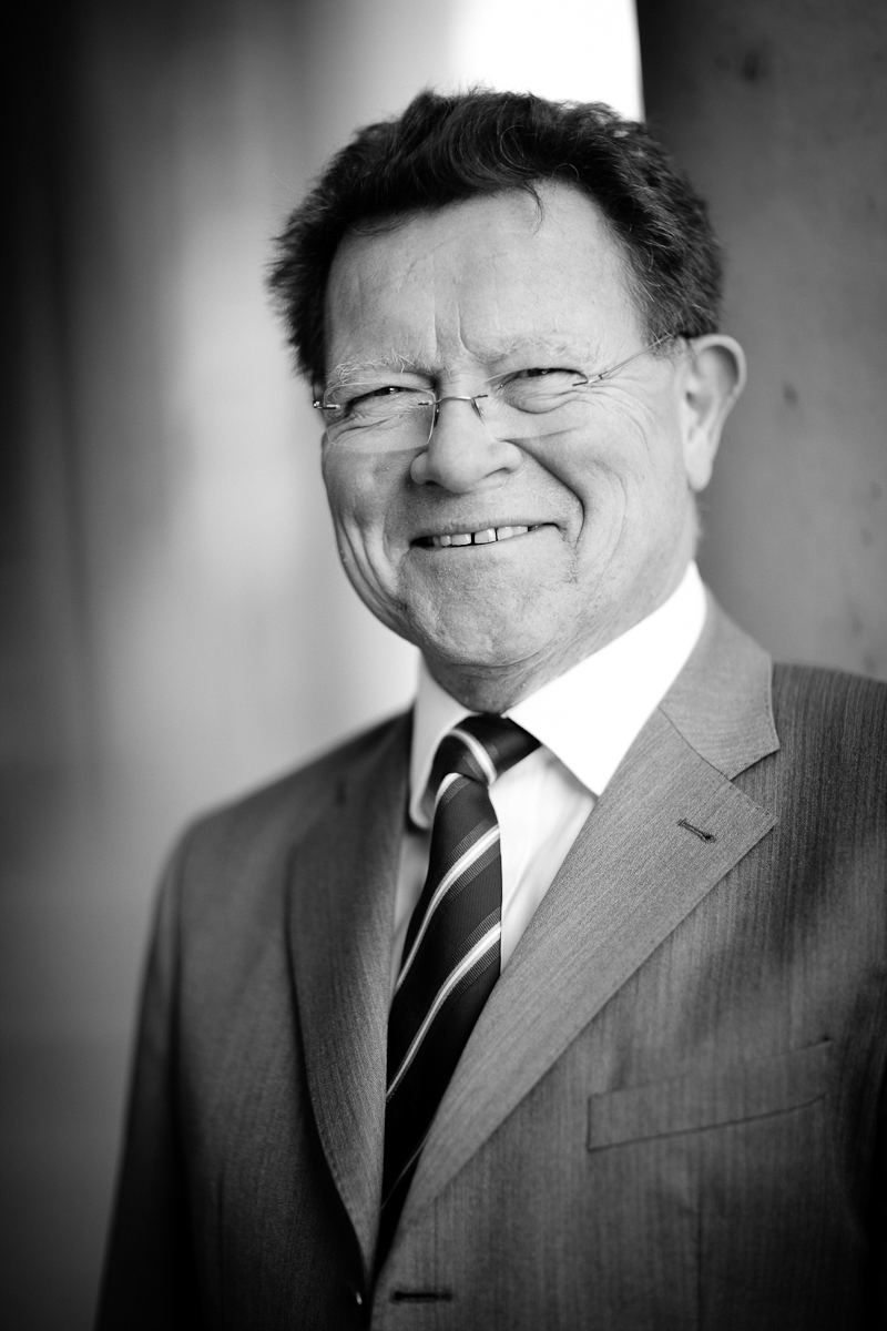 Ein lächelnder älterer Mann trägt einen Anzug und eine Krawatte, steht an einer Wand und schaut direkt in die Kamera. Sein Haar ist grau und er hat eine Brille auf. Das Bild ist in Schwarz-Weiß gehalten.