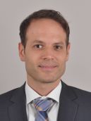 Porträt eines Mannes mit kurzen braunen Haaren, der in einem Anzug und einer Krawatte lächelt. Der Hintergrund ist neutral und unauffällig, um den Fokus auf die Person zu legen.