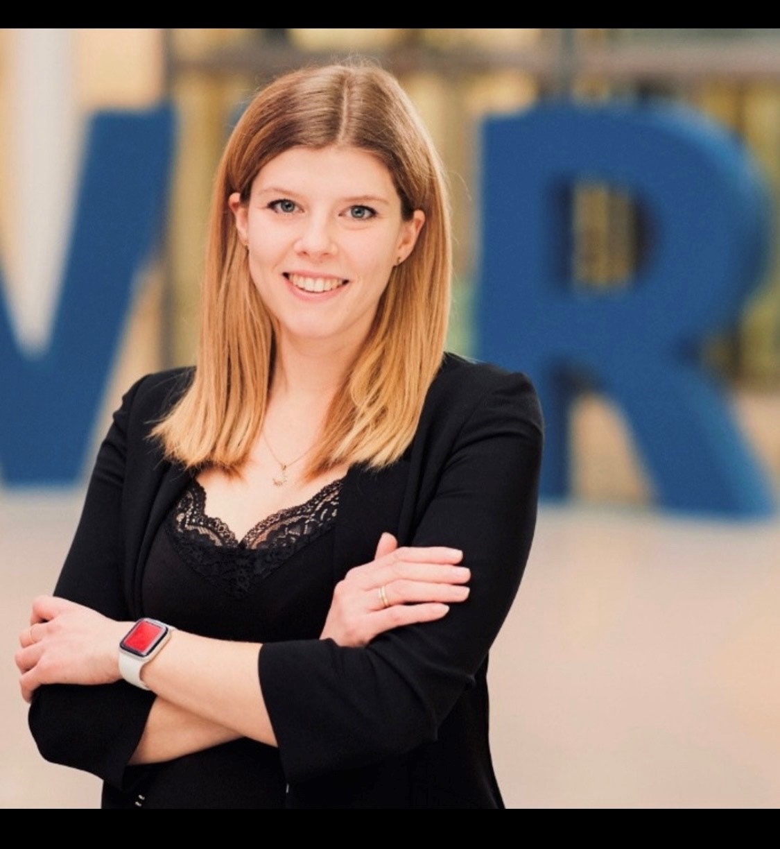 Eine lächelnde junge Frau mit blonden Haaren steht im Vordergrund, die Arme verschränkt. Sie trägt ein schwarzes Outfit und einen Fitness-Tracker am Handgelenk. Im Hintergrund sind große blaue Buchstaben zu sehen, die "VR" bilden.