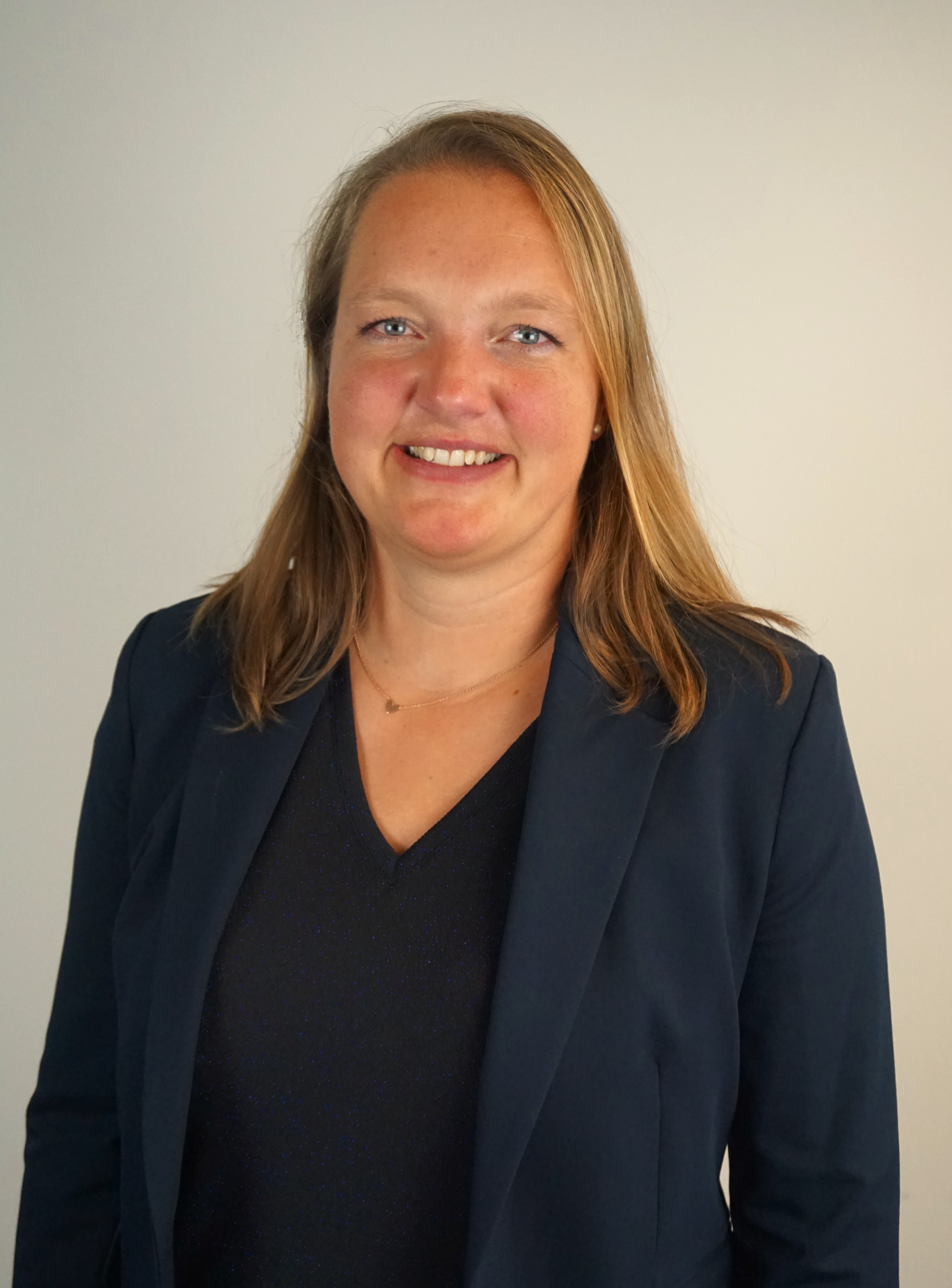 Eine Frau mit langen, blonden Haaren lächelt in die Kamera. Sie trägt einen dunklen Blazer über einem schwarzen Oberteil. Der Hintergrund ist hell und einfarbig, was ihre Präsenz betont.
