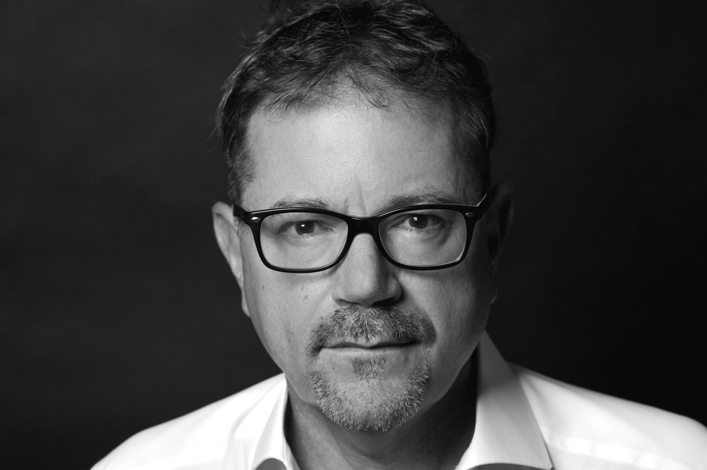 Ein Mann mit Brillengestell, kurzem Haar und einem leichten Bart schaut ernst in die Kamera. Er trägt ein weißes Hemd und hat einen neutralen Gesichtsausdruck. Der Hintergrund ist dunkel, was seine Gesichtszüge betont.